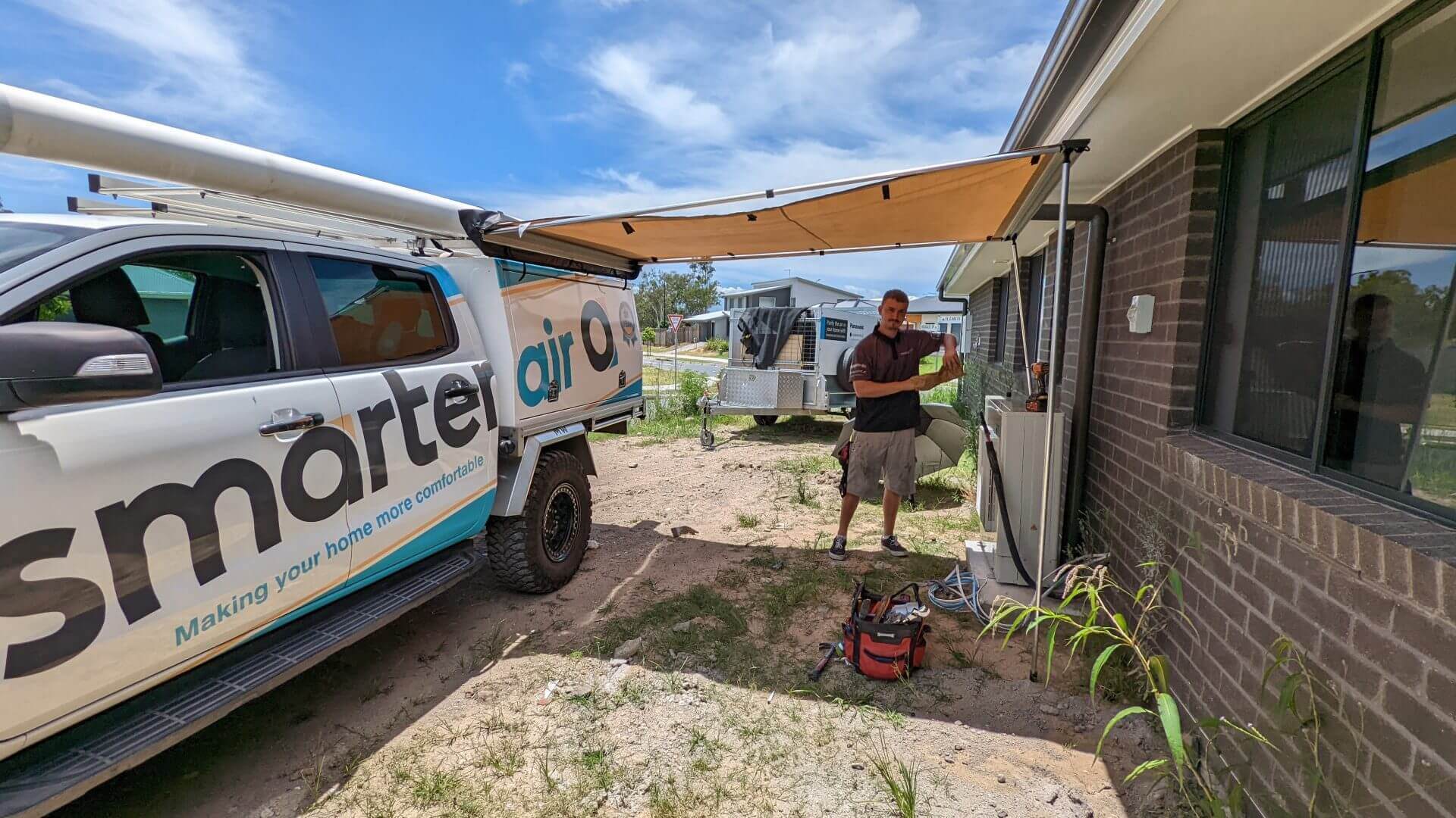 PXL_20211216_010931805 A technician stands beside a branded "Smarter Air" service vehicle with an awning extended, working on an outdoor air conditioning unit outside a brick house.