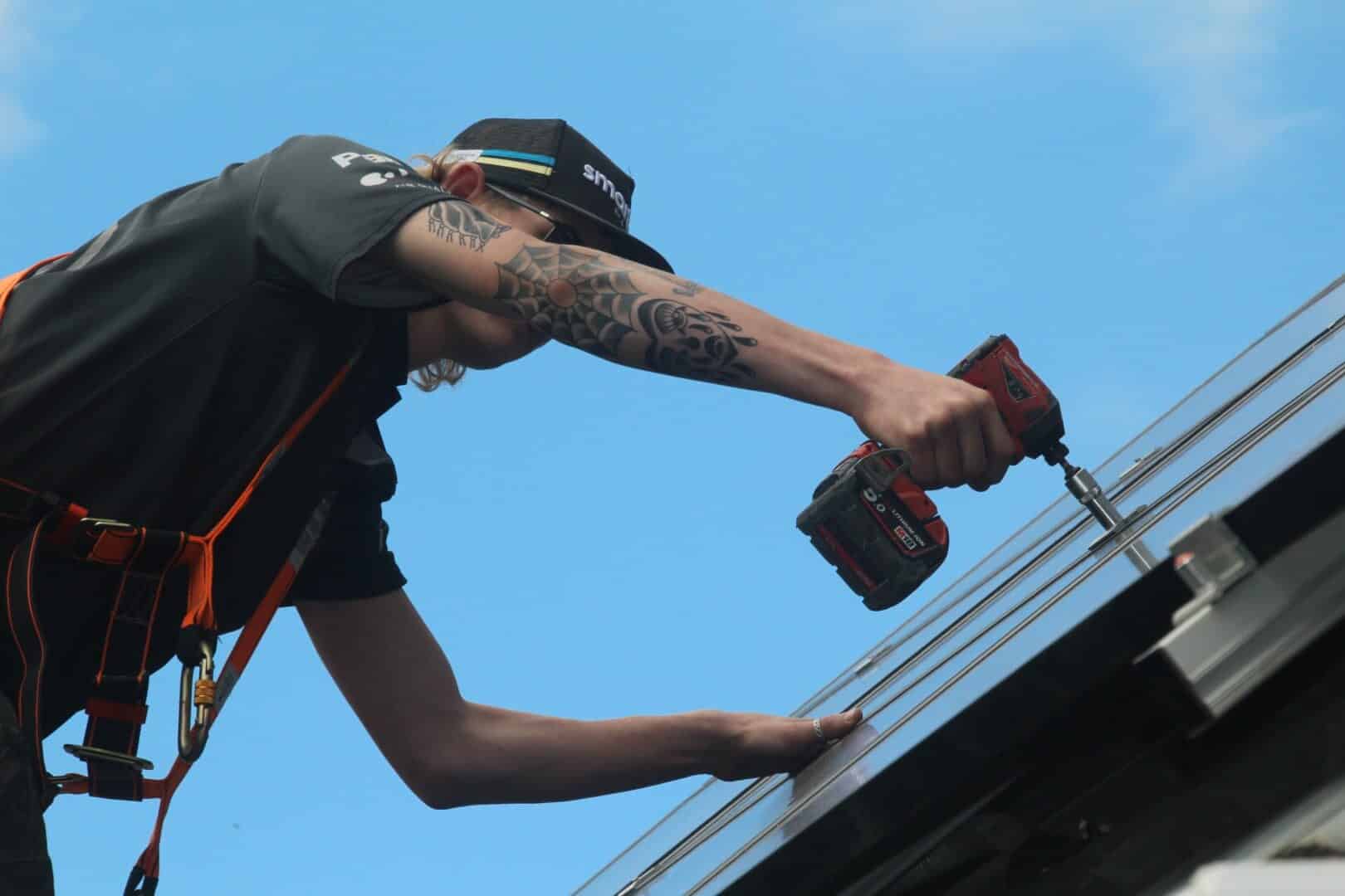 solar-panel-electrician-on-roof (10) A worker wearing a safety harness climbs a ladder on a solar-paneled roof, clipping onto a safety line while reaching toward the panels.
