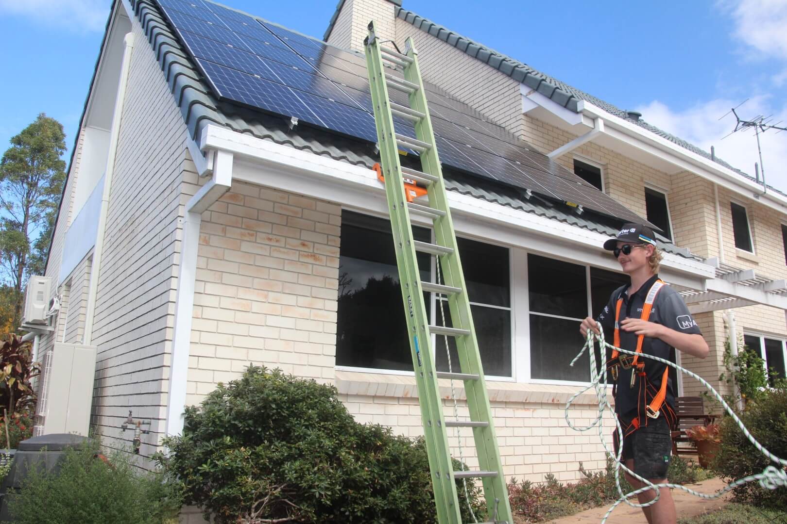 A worker wearing a safety harness stands beside a ladder leaning against a house with rooftop solar panels, holding a coiled safety rope.